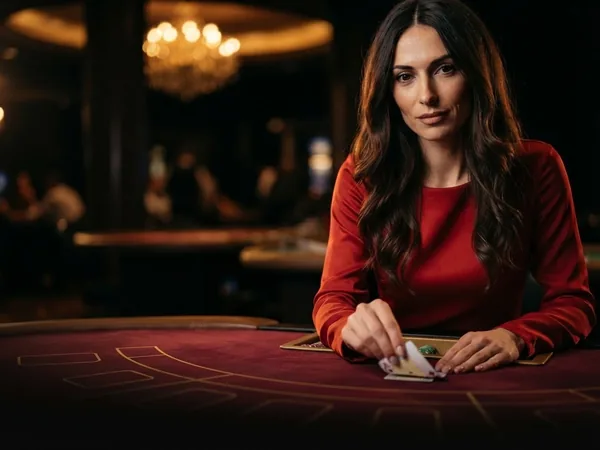 Lobby-style live table card: portrait crop of the croupier squeezing a card corner at a crimson table